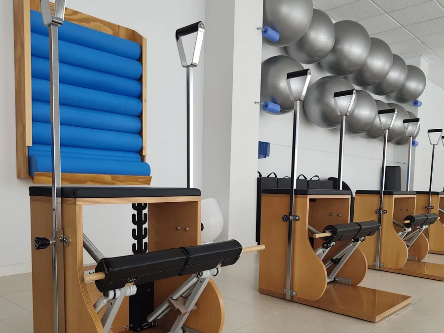 Primer plano de una silla de pilates de madera, con el panel de rulos azules y los fitballs detrás